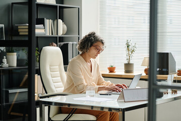 Poster - Side view of mature female employer typing on laptop keyboard while sitting by workplace in office and working with online data