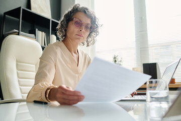 Poster - Focus on mature female chief executive officer reading financial paper while sitting by workplace in front of camera and checking data