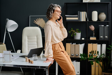 Poster - Smiling mature female boss with cellphone by ear calling business partner to arrange appointment or specify some organization points