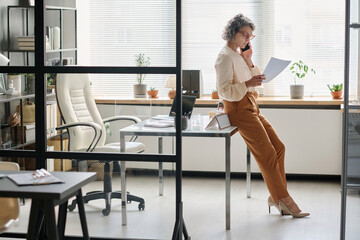 Poster - Side view of mature professional auditor or broker looking through contract terms or financial paper and talking on mobile phone