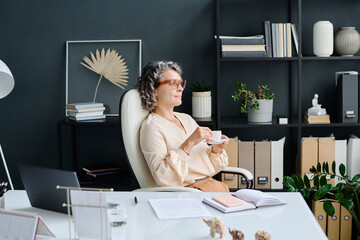 Wall Mural - Restful mature female ceo with cup of coffee sitting by workplace in office, looking through window and thinking of something pleasant