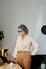 Poster - Mature businesswoman with grey hair touching keys of computer keyboard while standing by wooden desk with cup of coffee
