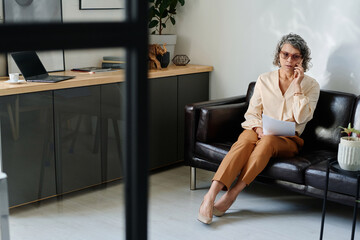 Poster - Mature female employer with resume of applicant making phone call while sitting on comfortable black leather couch and arranging appointment