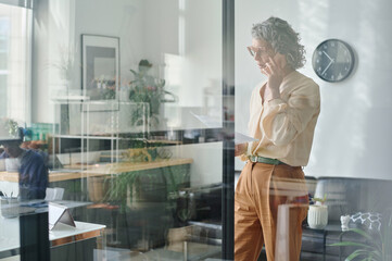 Poster - Confident mature businesswoman with paper document talking by smartphone to one of applicants while arranging appointment