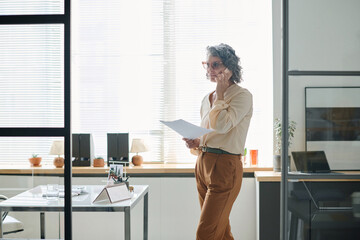 Poster - Confident mature businesswoman with cellphone by ear standing in spacious office and communicating with business partner or colleague