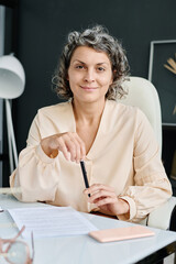 Poster - Successful mature entrepreneur or ceo with pen looking at camera with smile while sitting by workplace in office and working with documents