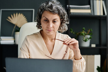 Poster - Serious mature female broker or analyst with eyeglasses in hand looking at laptop screen while sitting by workplace and analyzing financial data