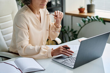 Wall Mural - Hand with pen of mature female analyst over laptop keyboard during network or work with online data by desk with open notebook