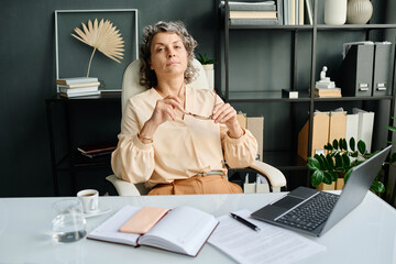 Poster - Confident mature female director of business company or corporation in quiet luxury apparel looking at camera while sitting by workplace