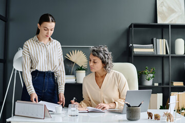 Poster - Young secretary or manager consulting with mature female boss about some points of contract or other business document in office