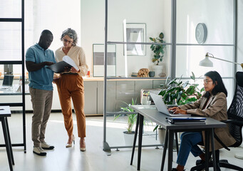 Aufkleber - Mature businesswoman and African American male employee discussing document while standing in the center of modern coworking area