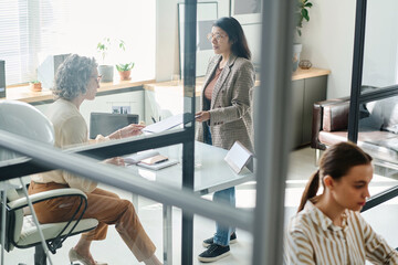 Poster - Young businesswoman passing financial document to mature female director of company while standing in front of her in spacious office