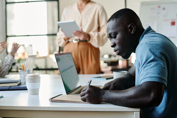 Leinwandbilder - Side view of young African American businessman making notes in notepad while sitting in front of camera during presentation