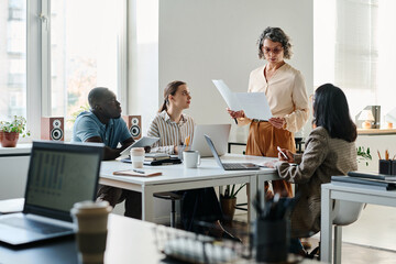 Wall Mural - Confident mature female director of business company with documents standing in front of several young intercultural white collar workers