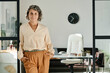 © pressmaster - Smiling mature female chief executive officer in beige blouse and brown pants standing in office against her workplace and looking at camera