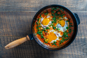 Wall Mural - Delicious shakshuka with eggs, tomato and parsley in a cast-iron pan, closeup, top view. Shakshuka - traditional israeli tomato stew with eggs