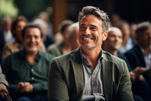 Smiling Man Sitting In Front Of Crowd Of People. This Image Can Be Used To Represent Leadership, Public Speaking, Or Confident Individual In Social Setting.