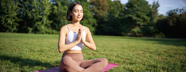 Wall Mural - Woman meditating on lawn in park, sitting on sports mat, relaxing, breathing fresh air