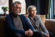 © 1by1step - A retired couple in casual clothes looking sad confused deep thought sit on sofa in living room