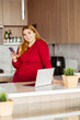 © Maria Vitkovska - Smiling beautiful plus size woman wearing red casual clothes holding mobile phone, using laptop