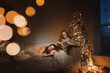 © illustrissima - Two teenage girls at home in the evening open gifts and read fairytales book in holidays christmas atmosphere sitting under new year tree