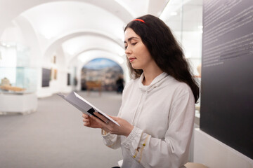 Side view of young caucasian woman student visiting museum exhibition and reading brochure. Light hall in background. Concept of cultural education and Museum's Day