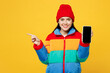 Young woman wear padded windbreaker jacket red hat casual clothes hold in hand use blank screen workspace area mobile cell phone chatting online point finger aside isolated on plain yellow background