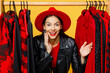 Young promoter surprised cheerful happy woman wearing red hat stand near rack of casual clothes looking camera scream isolated on plain yellow background. Black Friday shopping sale buy day concept.