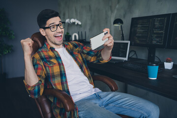 Portrait of crazy delighted web designer sit chair play video games smart phone raise fist triumph workplace business center indoors
