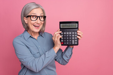 Photo of astonished positive woman with gray hairstyle dressed blue shirt in eyewear look at calculator isolated on pink background