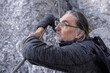 © Guzel - Adult gray-haired man sitting on stones, leaning on metal railing and looking into the distance, Austria
