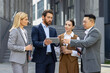 © Liubomir - Four business people discussing financial results of achievement, mature men and women outside office building discussing success strategy, diverse team of businessmen and businesswomen in suits