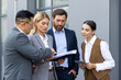 © Liubomir - Four business people discussing financial results of achievement, mature men and women outside office building discussing success strategy, diverse team of businessmen and businesswomen in suits
