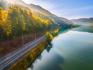 Wall Mural - aerial view of railroad near river in alpine mountains in fog at sunrise in autumn. top view of rura