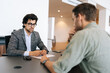 © dikushin - Selective focus of broker advisor in eyeglasses and business suit consulting to young man client sitting at desk. Professional male insurer talking consumer guy about insurance benefits at office.