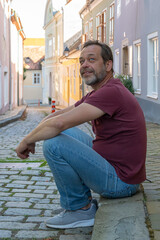 Wall Mural - An elderly man sits on the pavement on a narrow European pedestrian street and smiles.