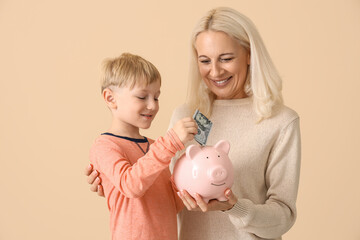 Wall Mural - Little boy with his grandmother putting money in piggy bank on beige background