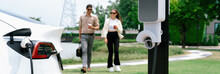 Young couple with coffee recharge EV car battery at charging station connect to electrical industrial power grid. Couple with shopping and travel using eco electric car lifestyle.Panorama Expedient