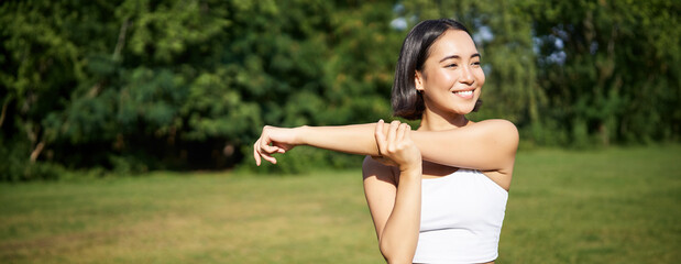 Wall Mural - Portrait of young fitness woman stretching her arms, warm-up before training session, sport event in park, jogging and excercising