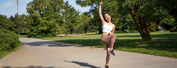 Wall Mural - Excited young asian woman winning, finish running in park, saying yes, lifting hand up in triumph, celebrating victory or success