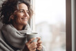 © Jasmina - Portrait of happy middle aged woman in cozy sweater holding a cup of hot drink and looking trough the window, enjoying the winter morning at home, side view