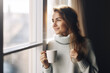© Jasmina - Portrait of happy young woman in cozy sweater holding a cup of hot drink and looking trough the window, enjoying the winter morning at home, side view