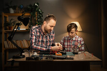 Father And His Son Using Soldering Iron And Multimeter For Fixing Motherboard From Laptop. Two Men Sitting Together At Table And Repairing Computer At Home.