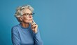 © Caphira Lescante - Senior woman with a finger tip on her cheek, thinking, happy with herself and her life. Best-ager, baby-boomer with grey short hair on a blue background.