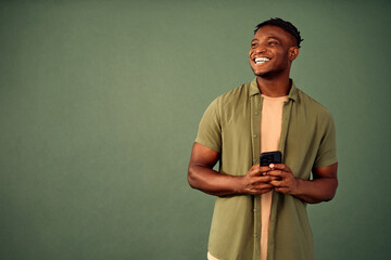 Wall Mural - New mobile app. Cheerful young man with toothy smile on face looking aside and using modern smartphone. African american guy in casual clothes isolated over green background with copy space.