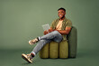 Modern lifestyles with gadgets. Cheerful guy in casual attire sitting on design pouf chair and holding digital tablet over green background. Smiling african man surfing internet or using new app.