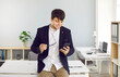 © Studio Romantic - Satisfied young businessman using mobile phone by phone near workplace in his office. Smiling man with glasses is using mobile phone for messaging, browsing websites or searching for e-commerce offers