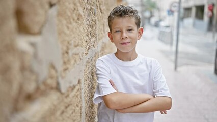 Poster - Adorable blond boy, confidently smiling, enjoys fun standing in an urban street, positively expressing joy with his arms crossed, leaning against a city-wall outdoors