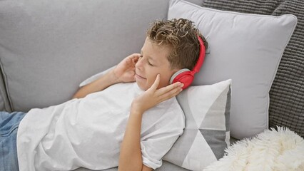Wall Mural - Adorable blond child, smiling with confidence, engrossed in music, lying restful on sofa, enjoying audio device tech at home