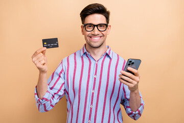 Sticker - Photo of shopaholic geek man holding plastic credit card using smartphone for online shopping website isolated on beige color background